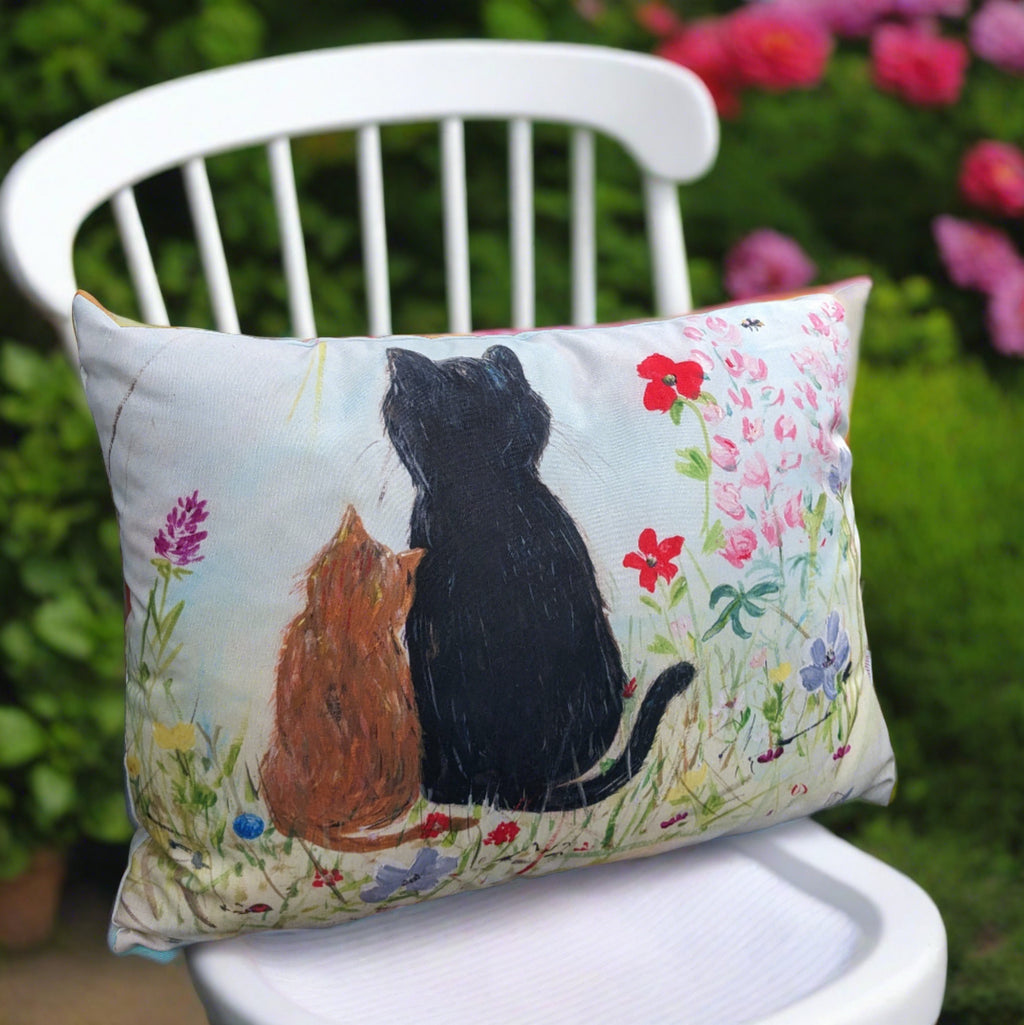 outdoor cushion. features a Ginger kitten resting it's head on a black cat in a garden